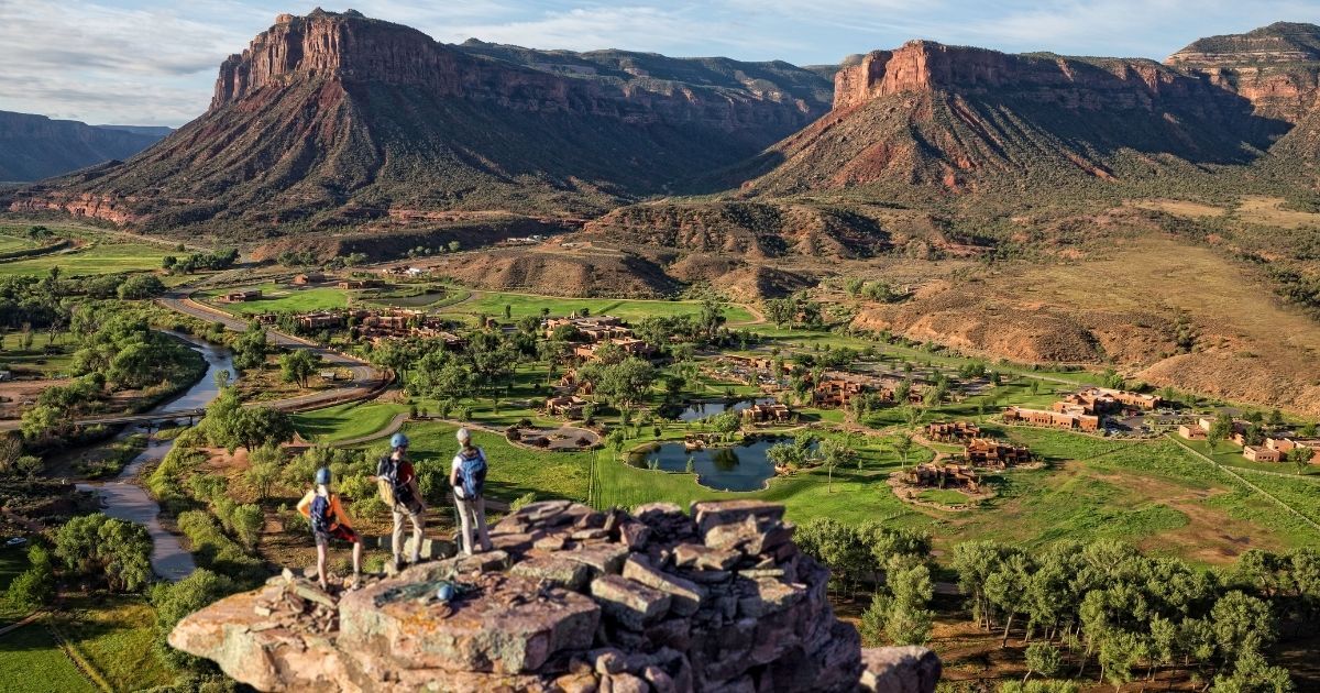 Three people stand on a rocky outcrop, overlooking a green valley with scattered buildings, trees, and a river. In the background, there are red rock formations and a blue sky.