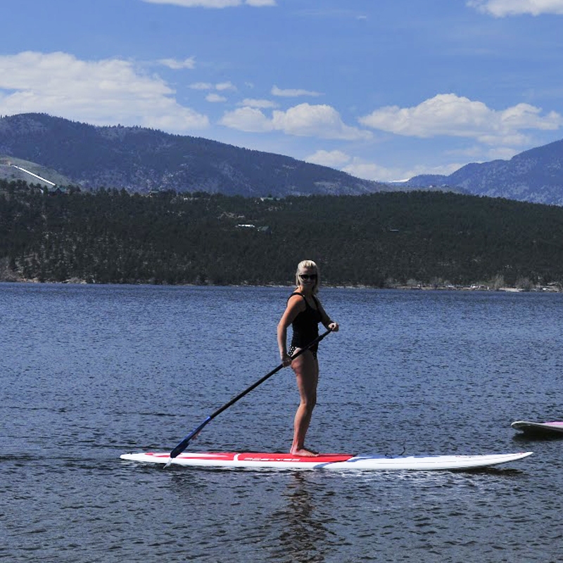 A person stands on a paddleboard in a lake with mountainous terrain in the background, holding a paddle on a sunny day.