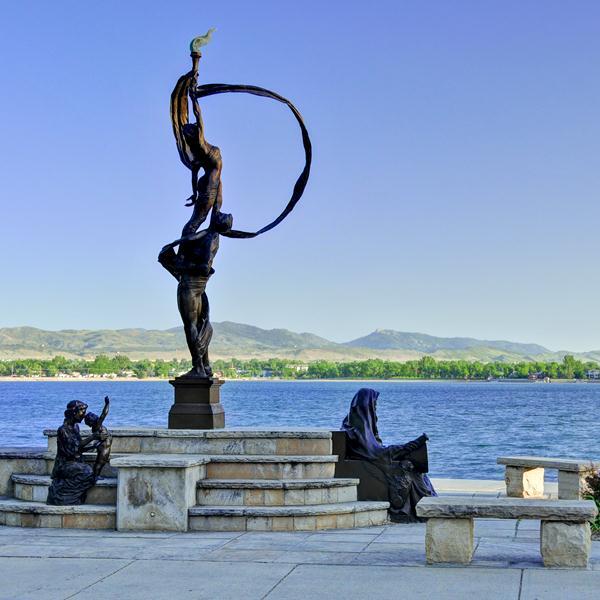 A bronze sculpture of a man holding a circular ribbon with a bird atop stands on a pedestal by a lake, accompanied by seated figures on the surrounding stone benches and steps.