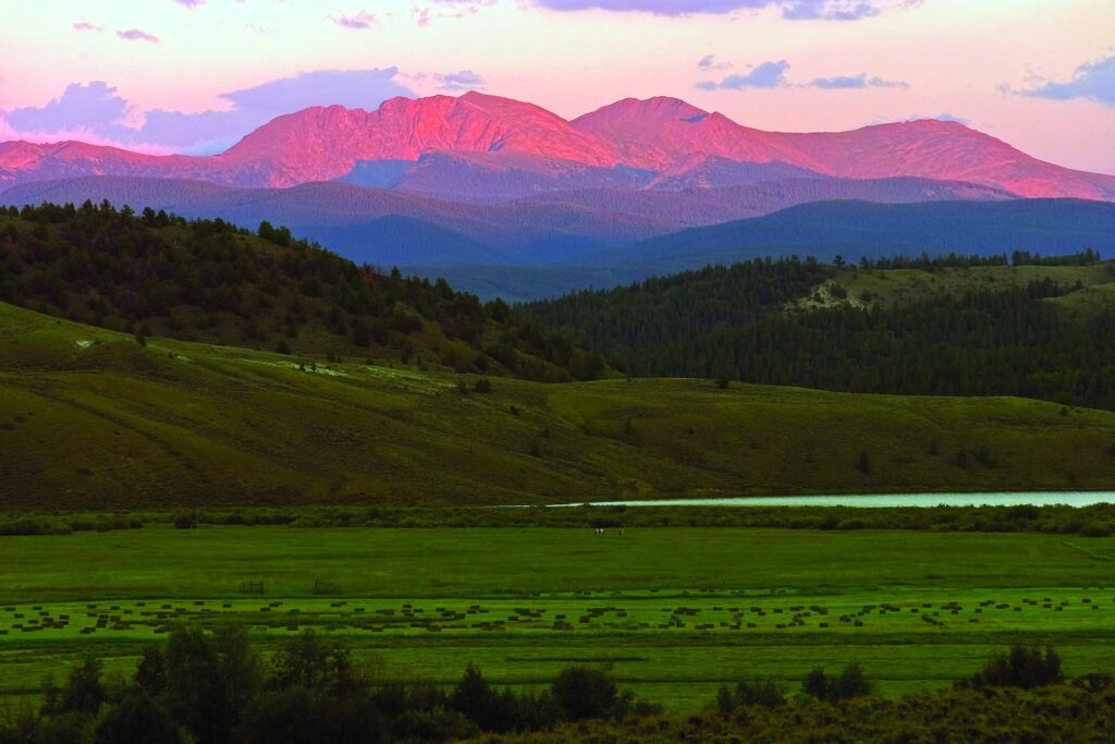Sunset over mountainous landscape with rolling hills, a green valley, and a partially visible lake. The mountain peaks are tinted pink and orange by the setting sun.