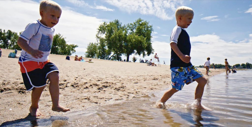 Two young children happily splash in shallow water at a sunny beach, with trees and people in the background.