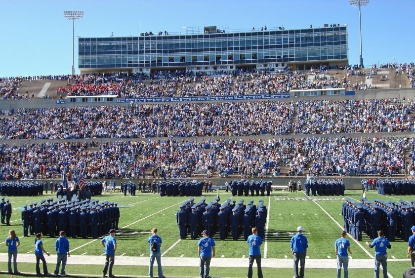 A large formation of uniformed personnel on a football field with a backdrop of filled stadium stands. Spectators watch the organized assembly under a clear blue sky.