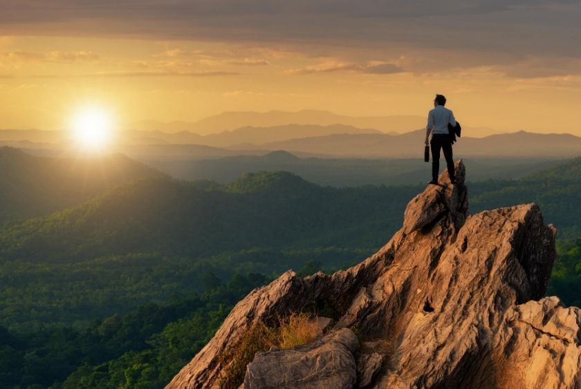 A person stands on a rocky peak at sunset, overlooking a vast landscape of forests and distant hills.
