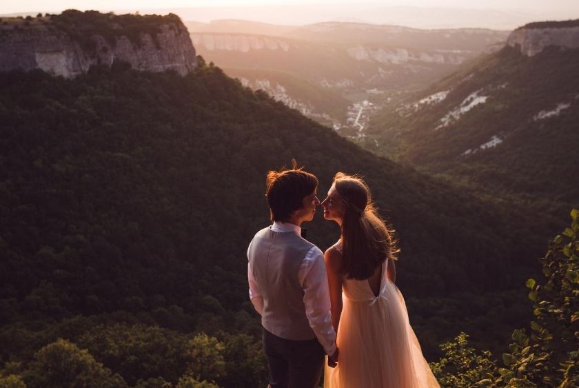 A couple stands on a mountainside at sunset, facing each other and holding hands, with a scenic valley and cliffs in the background—a perfect moment for an unforgettable destination wedding.