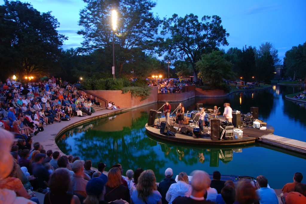 Outdoor evening concert with a small band performing on a circular stage above water, surrounded by a large audience seated on steps and grass. Trees and evening sky in the background.