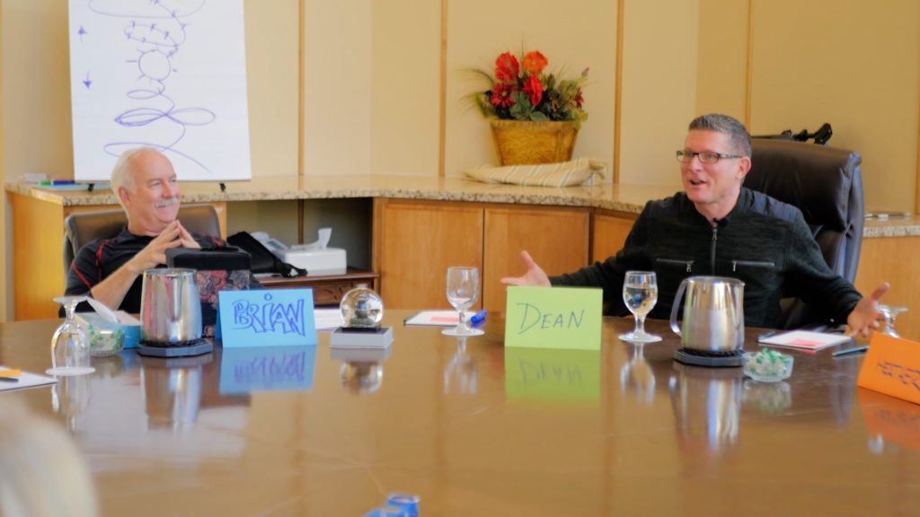 Two men are seated at a conference table with name placards labeled 