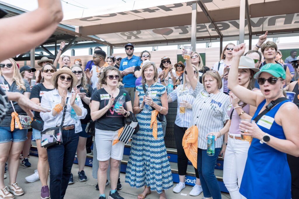 A group of people gathered outdoors wearing casual attire, some holding drinks and raising their hands. They are standing in front of a shaded structure with people sitting in the background.