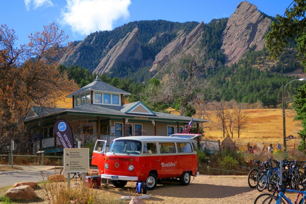 A red Volkswagen van is parked near a visitor center with a wooden house in a mountainous, autumnal landscape. Several bicycles are seen on the right side of the image.