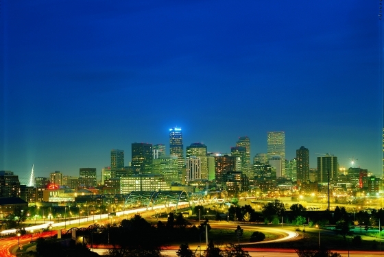 A nighttime view of a city skyline with buildings illuminated against a clear blue sky, and light trails from vehicles on a highway in the foreground.