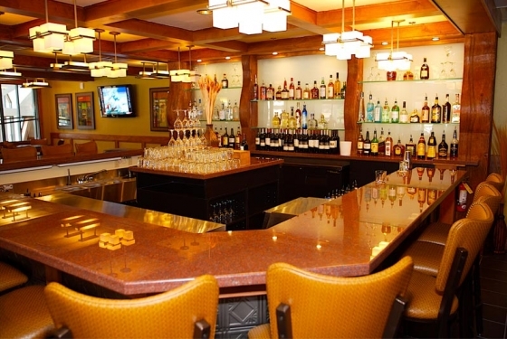 A well-lit bar with a wooden counter, chairs, and shelves stocked with various bottles of liquor. Glasses are neatly arranged on the counter, and a TV is mounted on the wall in the background.