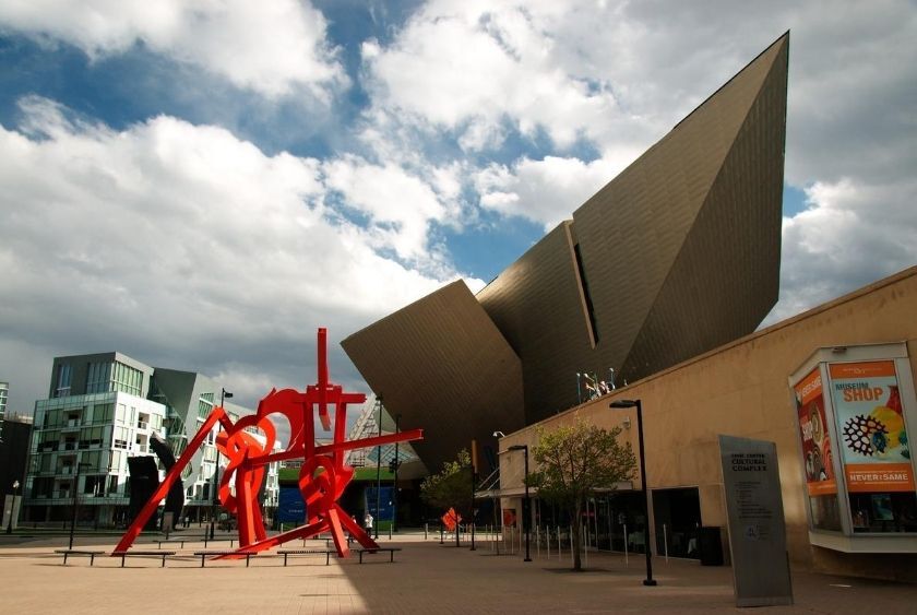 Modern architectural building with sharp angles, a large red abstract sculpture, informational sign, and cloudy sky in the background.