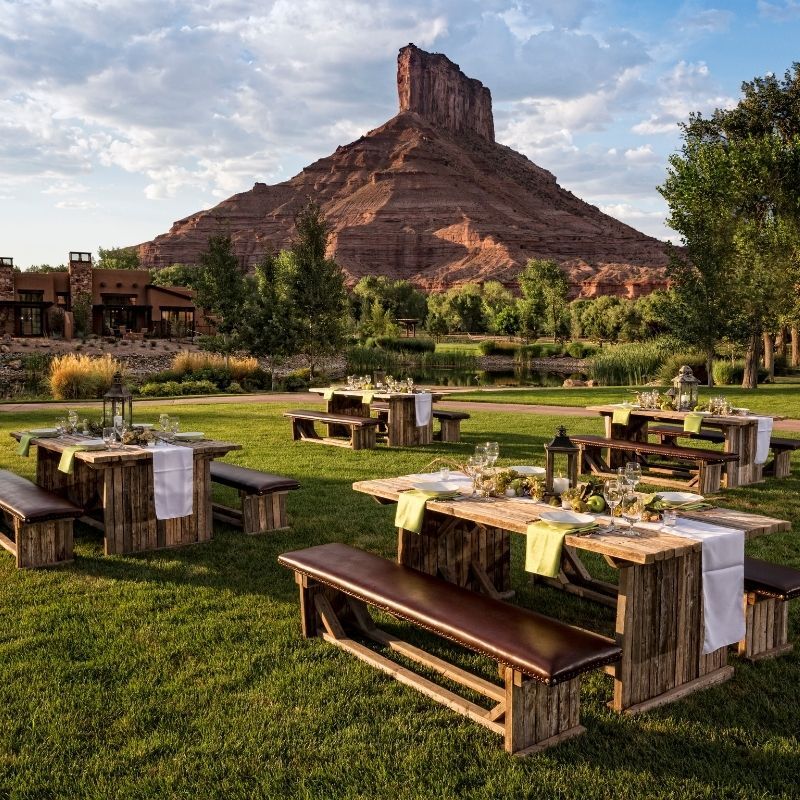 Outdoor dining tables and wooden benches set up on a grassy lawn, with a rock formation and trees in the background under a partly cloudy sky.