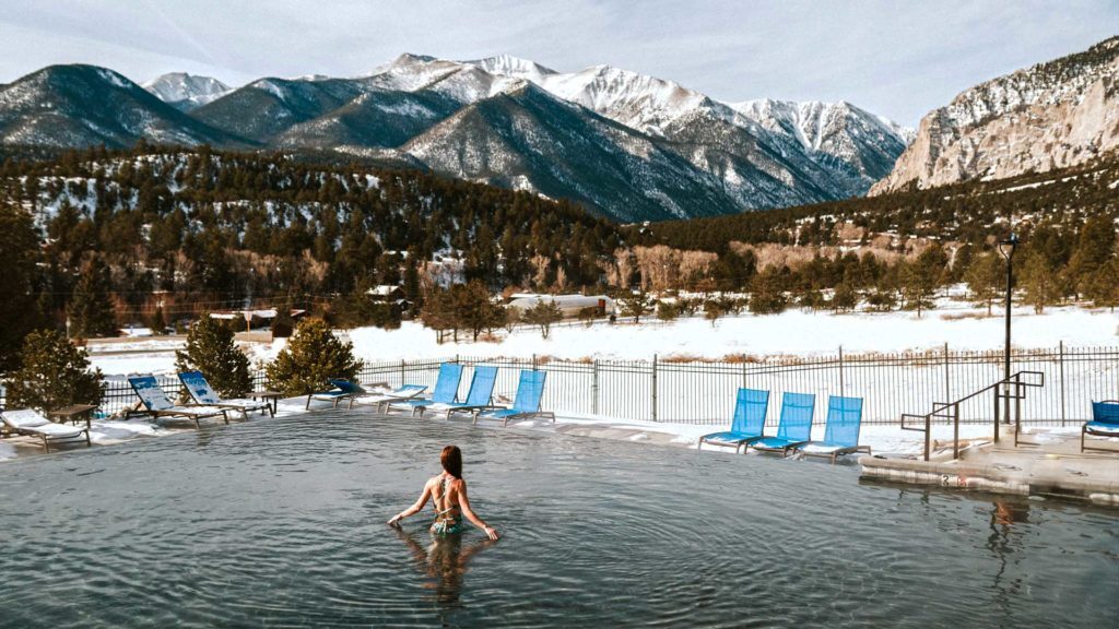 A person in a swimsuit stands in an outdoor pool with snowy mountains and trees in the background. Several empty lounge chairs are arranged around the pool.
