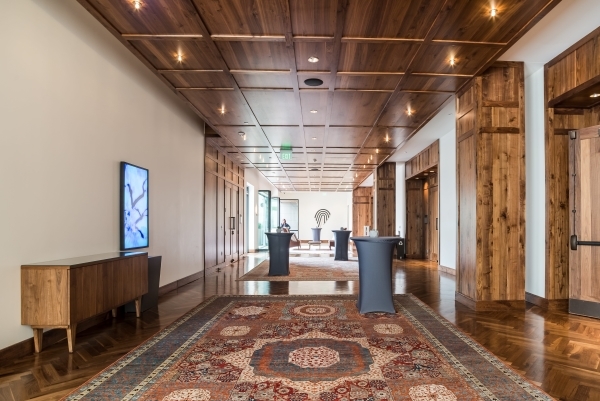 A modern hallway with wooden walls and ceiling, a patterned rug on the floor, a console table on the left, and two high tables with dark coverings. Bright lighting illuminates the space.