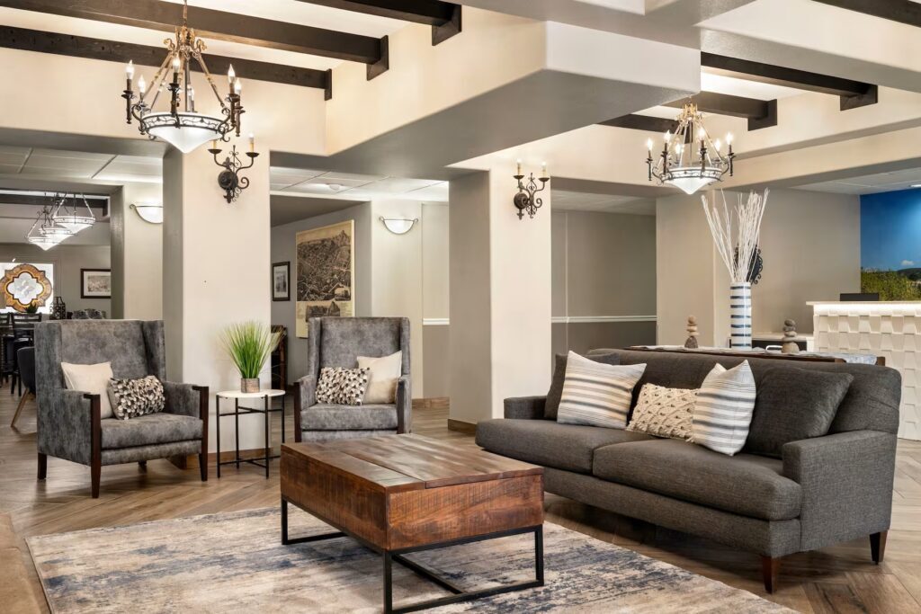 Modern living room with gray sofas and armchairs, a wooden coffee table, and two chandeliers hanging from a beamed ceiling. The room has light-colored walls with sconce lighting and decorative plants.