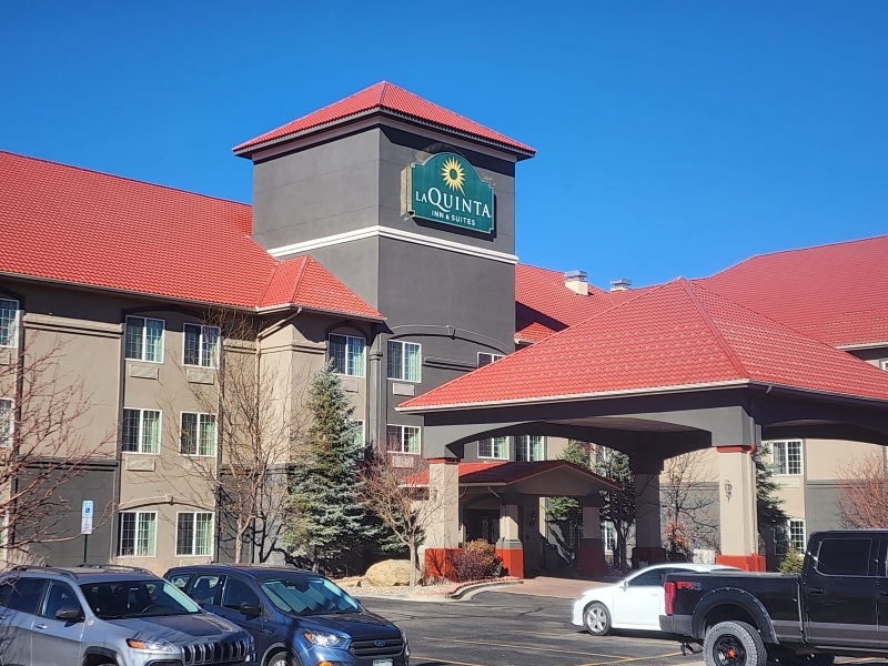 Exterior of a La Quinta Inn & Suites hotel building with a red roof and a light brown facade. There are several parked cars in front of the entrance. The sky is clear and blue.