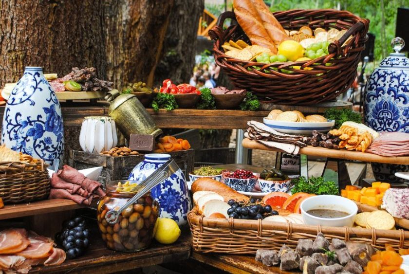 A rustic outdoor spread featuring various breads, cheeses, olives, fruits, meats, and decorative blue and white vases, all arranged on wooden tables with greenery accents.