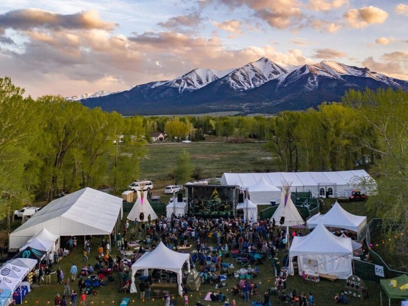 A crowd gathers at an outdoor festival with white tents and tipis, set against a backdrop of snow-capped mountains and green trees under a partly cloudy sky.