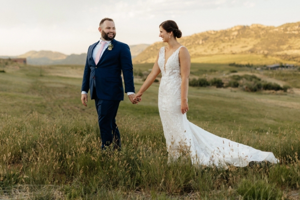 A bride and groom hold hands while walking through a grassy field with mountains in the background. The bride is wearing a white dress, and the groom is in a blue suit.