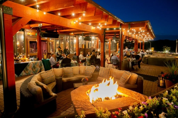 Outdoor restaurant seating area with a fire pit in the center, surrounded by cushioned seats and string lights overhead. Patrons are dining at tables under a wooden canopy with warm, ambient lighting.